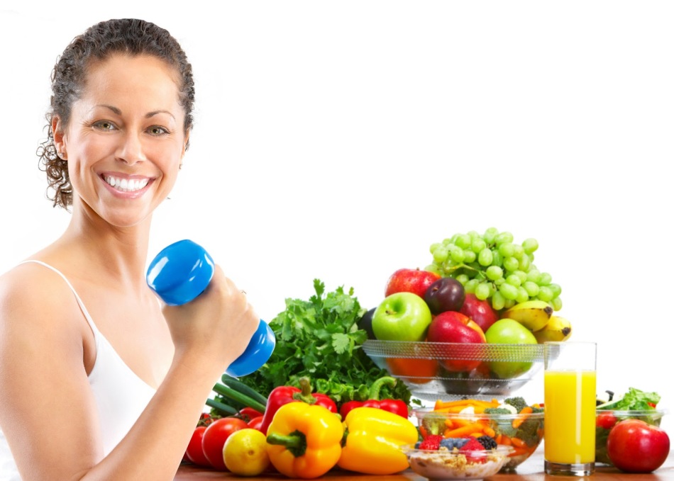 Улыбчивая брюнетка с гантелей в руке на фоне овощей и фруктов, которые можно кушать после тренировки